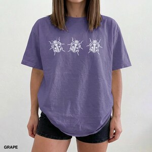 May include: A grape-colored short-sleeve t-shirt featuring a white graphic print of three ladybugs arranged in a horizontal row across the chest. The shirt has a crew neckline and a relaxed fit, showcasing a simple and clean design aesthetic.