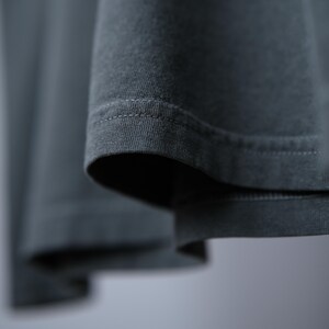 May include: A dark charcoal gray garment featuring a visible hem with double-stitched detailing. The fabric has a soft, matte texture and is captured in a close-up view that highlights the clean, horizontal lines of the stitching along the edge.