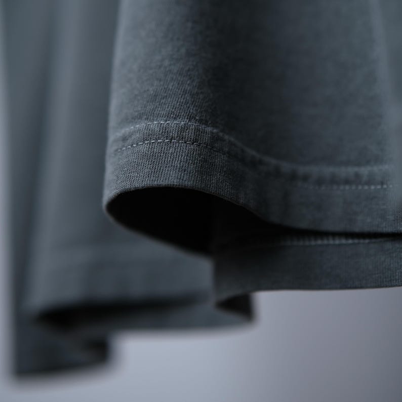 May include: A dark charcoal gray garment featuring a visible hem with double-stitched detailing. The fabric has a soft, matte texture and is captured in a close-up view that highlights the clean, horizontal lines of the stitching along the edge.