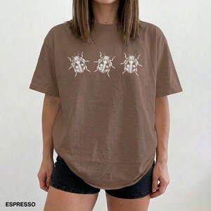 May include: An espresso brown short-sleeved t-shirt featuring a horizontal row of three white line-art illustrations of beetles centered on the chest. The shirt has a crew neckline and a relaxed fit, with the word ESPRESSO printed in the bottom corner.