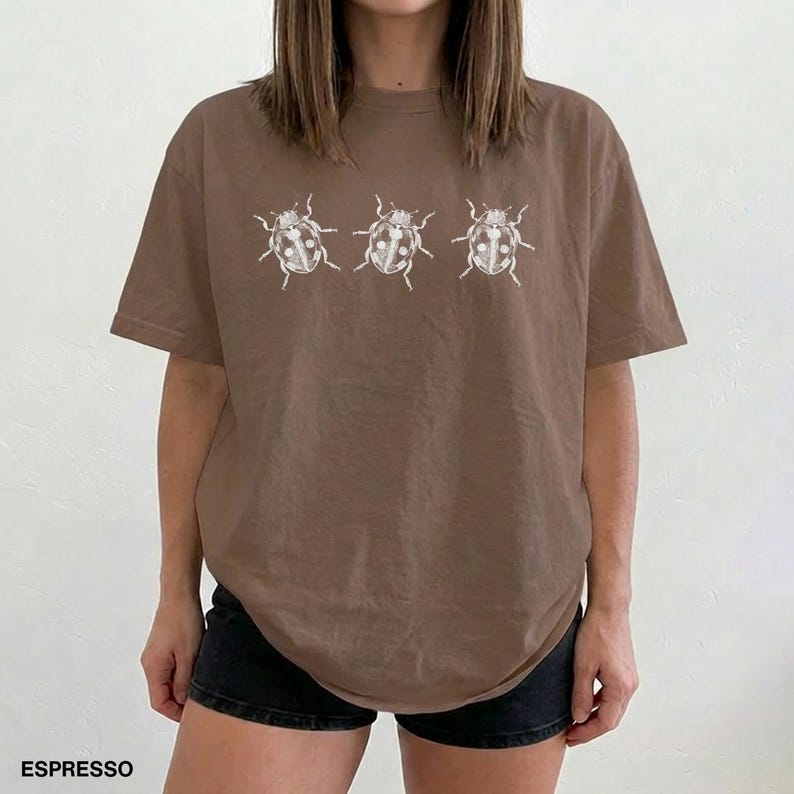 May include: An espresso brown short-sleeved t-shirt featuring a horizontal row of three white line-art illustrations of beetles centered on the chest. The shirt has a crew neckline and a relaxed fit, with the word ESPRESSO printed in the bottom corner.