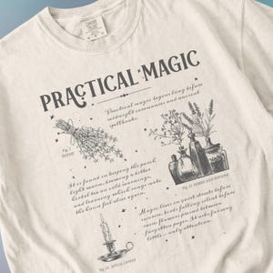 May include: Cream-colored sweatshirt with the words "PRACTICAL MAGIC" in dark gray. The design includes illustrations of thyme, herbs and potions, and a ritual candle. Text describes practical magic and its origins.