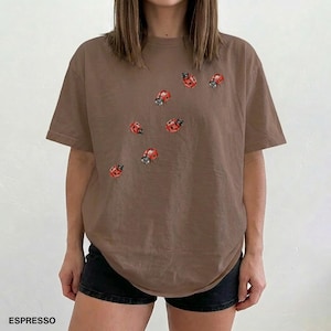 Może przedstawiać: Brązowy t-shirt z okrągłym dekoltem i kr&oacute;tkimi rękawami, ozdobiony motywami czerwonych biedronek. Słowo "ESPRESSO" jest wydrukowane na dole. Ten swobodny top jest odpowiedni do noszenia na co dzień. Koszulka jest w odcieniu brązu.