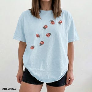 Może przedstawiać: Jasnoniebieski t-shirt z okrągłym dekoltem i kr&oacute;tkimi rękawami. Koszulka ma wz&oacute;r czerwonych i czarnych biedronek. Słowo "CHAMBRAY" jest wydrukowane na czarno w lewym dolnym rogu obrazu.