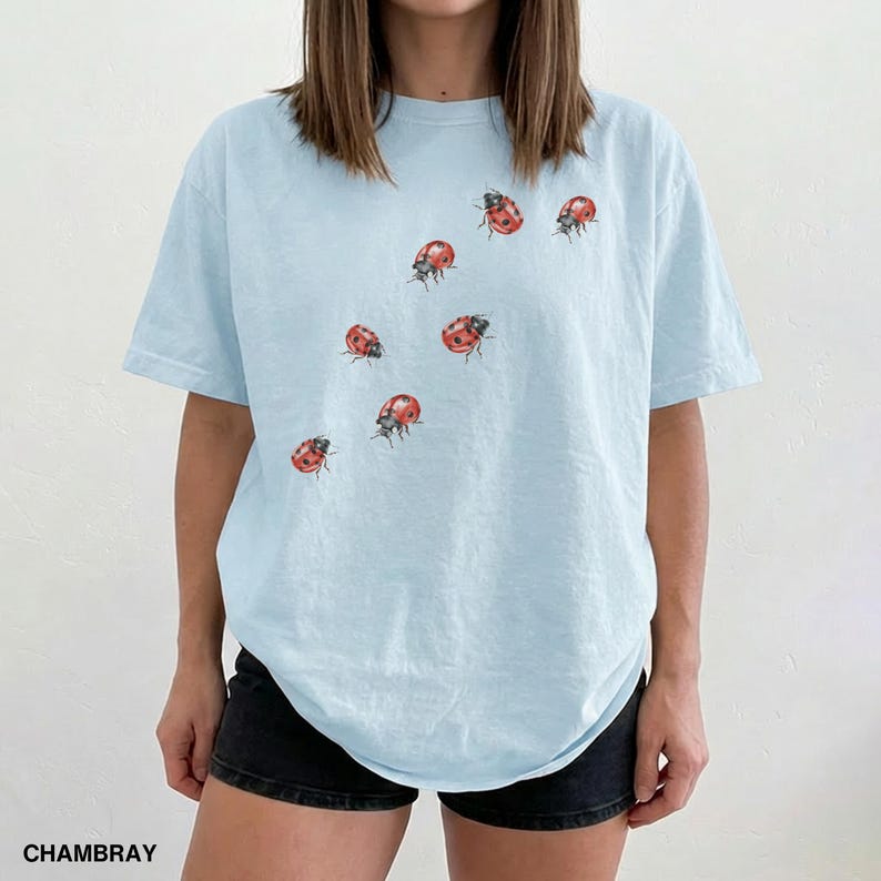 Może przedstawiać: Jasnoniebieski t-shirt z okrągłym dekoltem i kr&oacute;tkimi rękawami. Koszulka ma wz&oacute;r czerwonych i czarnych biedronek. Słowo "CHAMBRAY" jest wydrukowane na czarno w lewym dolnym rogu obrazu.