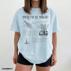 May include: Light blue t-shirt with the text "PRACTICAL MAGIC" and botanical illustrations. The shirt features drawings of herbs, bottles, and a candle, with accompanying text. The shirt is worn with black shorts.