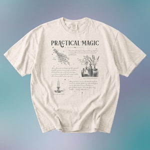 May include: Cream-colored t-shirt with the words "PRACTICAL MAGIC" in a stylized font. The shirt features illustrations of herbs, bottles, and a candle, along with handwritten text. The design is printed in dark gray.