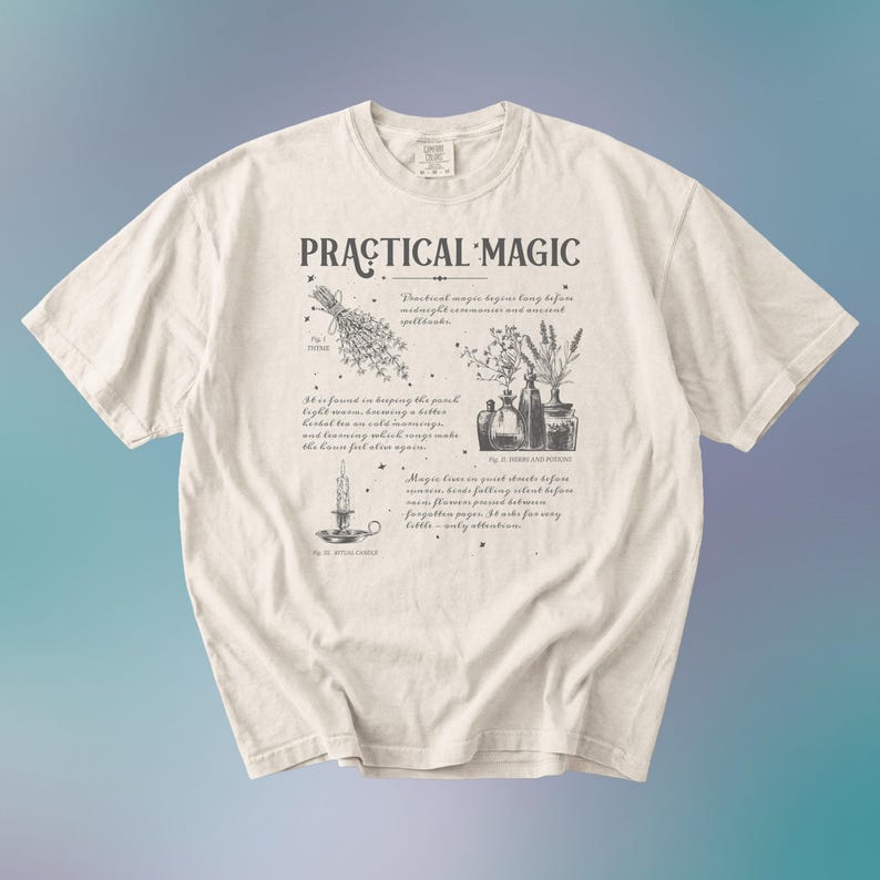 May include: Cream-colored t-shirt with the words "PRACTICAL MAGIC" in a stylized font. The shirt features illustrations of herbs, bottles, and a candle, along with handwritten text. The design is printed in dark gray.
