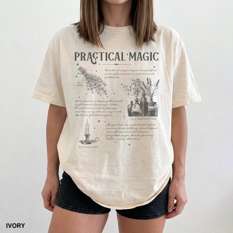 May include: Ivory-colored t-shirt with the words "PRACTICAL MAGIC" in a vintage font. The shirt features a detailed graphic with botanical illustrations and text, including the word "IVORY" at the bottom. A stylish and unique tee.