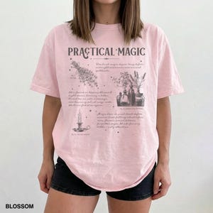 May include: A light pink t-shirt with the words "PRACTICAL MAGIC" in a stylized font. The shirt features illustrations of herbs, candles, and potion bottles. The word "BLOSSOM" is printed at the bottom left.