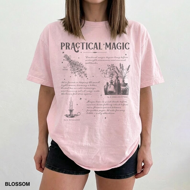 May include: A light pink t-shirt with the words "PRACTICAL MAGIC" in a stylized font. The shirt features illustrations of herbs, candles, and potion bottles. The word "BLOSSOM" is printed at the bottom left.