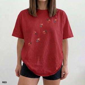 Może przedstawiać: Czerwony t-shirt z okrągłym dekoltem i kr&oacute;tkimi rękawami. Koszulka posiada liczne haftowane wzory biedronek. Słowo "RED" jest wydrukowane w lewym dolnym rogu. Modelka ma na sobie czarne szorty.