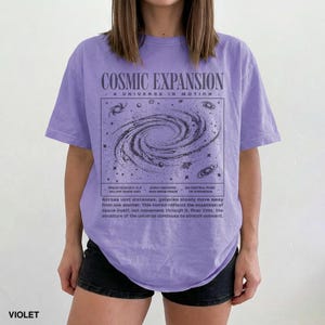 May include: Violet t-shirt with a graphic of a galaxy and the words "COSMIC EXPANSION" and "A UNIVERSE IN MOTION". The graphic includes text about the universe's expansion. The shirt is a relaxed fit.