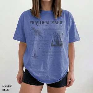 May include: Mystic blue t-shirt with the words "PRACTICAL MAGIC" printed on the front. The design includes illustrations of herbs, bottles, and a candle. The shirt is a relaxed fit.