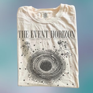Puede incluir: Una camiseta de color crema con un estampado gráfico negro de un agujero negro en espiral rodeado por dos astronautas y estrellas. El texto THE EVENT HORIZON está impreso en una fuente serif negrita sobre el gráfico. Cuello redondo.