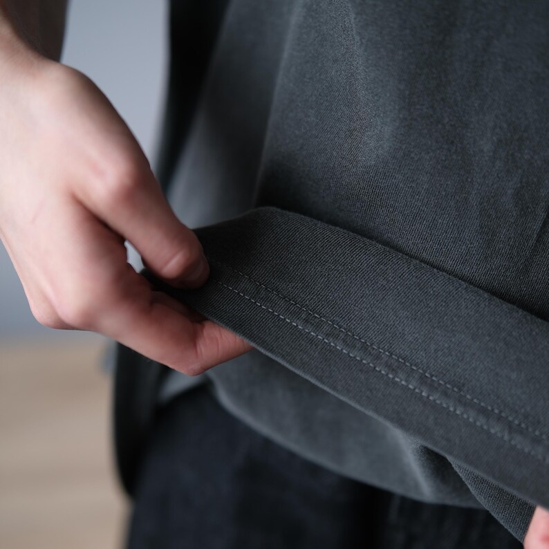May include: A dark charcoal gray t-shirt featuring a short sleeve with a folded hem. The fabric has a matte, slightly washed texture with visible double-needle stitching along the bottom edge of the sleeve cuff, showcasing the garment construction.