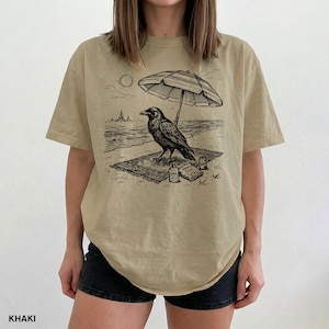 May include: Khaki-colored t-shirt featuring a black and white illustration of a raven relaxing on a beach under an umbrella. The raven is on a rug with a bottle and a snack. The word "KHAKI" is printed at the bottom.