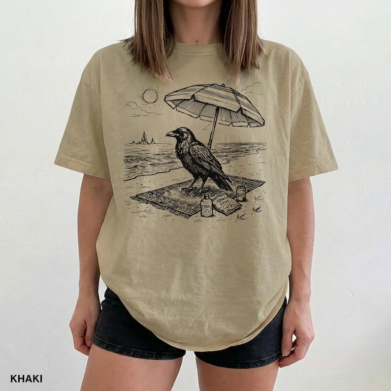 May include: Khaki-colored t-shirt featuring a black and white illustration of a raven relaxing on a beach under an umbrella. The raven is on a rug with a bottle and a snack. The word "KHAKI" is printed at the bottom.