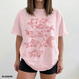 May include: A light pink short-sleeve t-shirt featuring a graphic print of multiple goldfish in a red line-art style. The design includes various fish swimming in different directions across the front of the garment. The word BLOSSOM is printed in the bottom left corner.