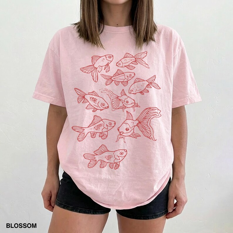 May include: A light pink short-sleeve t-shirt featuring a graphic print of multiple goldfish in a red line-art style. The design includes various fish swimming in different directions across the front of the garment. The word BLOSSOM is printed in the bottom left corner.