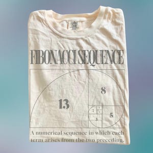 Könnte beinhalten: Cremefarbenes T-Shirt mit der Aufschrift "FIBONACCI SEQUENCE" und einer Darstellung der Fibonacci-Folge. Die Zahlen 1, 2, 3, 5, 8 und 13 sind sichtbar. Der Text unten lautet: "Eine Zahlenfolge, bei der sich jedes Glied aus den beiden vorhergehenden ergibt."