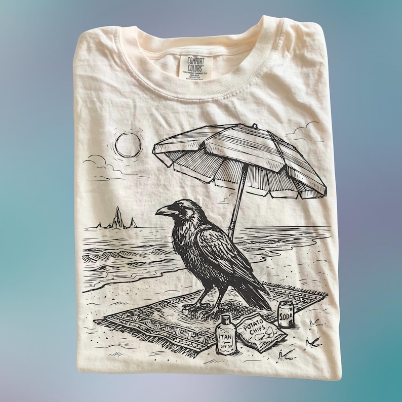 May include: Cream-colored t-shirt featuring a black and white illustration of a raven relaxing on a beach towel under a striped umbrella. The design includes a sun, ocean, and beach accessories like sunscreen, potato chips, and soda.