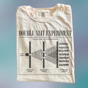 May include: A cream-colored t-shirt featuring a black graphic print of the double-slit experiment. The design includes the text "DOUBLE-SLIT EXPERIMENT" and "QUANTUM INTERFERENCE" with a diagram showing a light source, a single slit screen, a double slit screen, and an interference pattern.