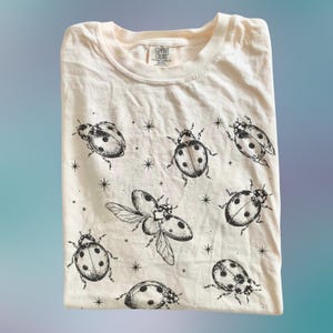 Puede incluir: Una camiseta de color crema con un estampado repetido en tinta negra de mariquitas en varias posiciones, incluyendo una con las alas extendidas, intercaladas con pequeñas estrellas de cuatro puntas. La etiqueta interior muestra el texto Comfort Colors.