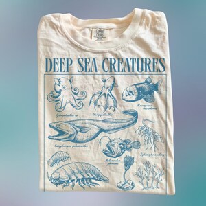 以下が含まれることがあります： クリーム色のTシャツ。青色のイラストで深海生物が描かれており、タコやチョウチンアンコウなどが含まれています。「DEEP SEA CREATURES」の文字がイラストの上にプリントされています。Tシャツは折りたたまれています。