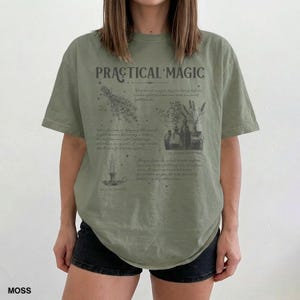 May include: Moss green t-shirt with the text "PRACTICAL MAGIC" and botanical illustrations. The shirt features a crew neck and short sleeves. The design includes images of herbs, bottles, and a candle.