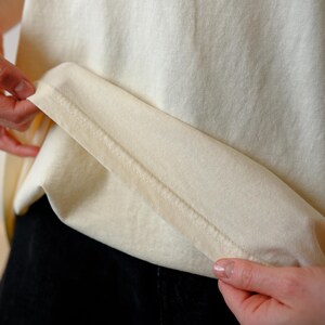 May include: A plain, off-white t-shirt featuring a visible hemline with double-stitched detailing along the bottom edge. The fabric has a smooth, matte texture and a light cream color, showcasing the construction of the garment's lower hem.