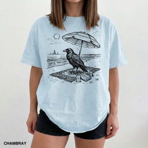 May include: Light blue t-shirt featuring a black and white illustration of a raven on a beach. The raven is under a striped umbrella, with the sun and ocean in the background. The shirt also has the word "CHAMBRAY" printed at the bottom.