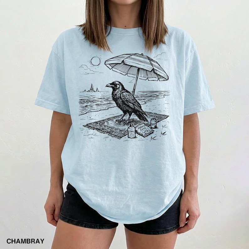 May include: Light blue t-shirt featuring a black and white illustration of a raven on a beach. The raven is under a striped umbrella, with the sun and ocean in the background. The shirt also has the word "CHAMBRAY" printed at the bottom.
