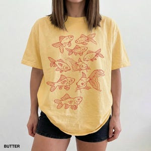 May include: A butter yellow short-sleeve t-shirt featuring a graphic print of multiple goldfish in a reddish-orange ink. The design consists of several fish arranged in a cluster across the front of the garment, showcasing various swimming positions.