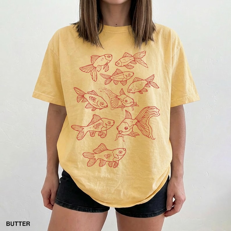 May include: A butter yellow short-sleeve t-shirt featuring a graphic print of multiple goldfish in a reddish-orange ink. The design consists of several fish arranged in a cluster across the front of the garment, showcasing various swimming positions.