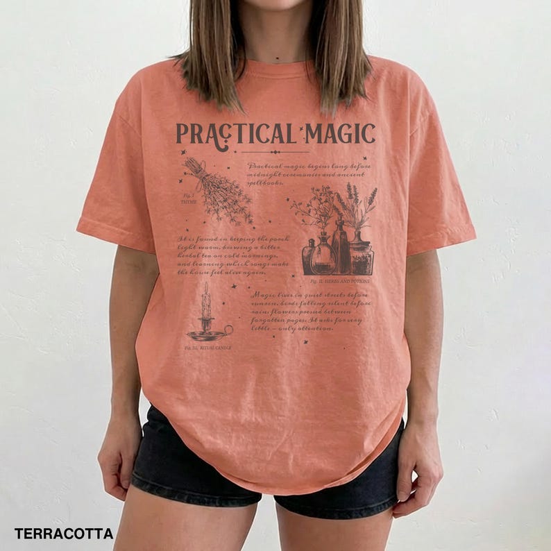 May include: Terracotta-colored t-shirt with the words "PRACTICAL MAGIC" printed on the front. The shirt features illustrations of herbs, bottles, and a candle. The text below the graphic describes the practice of magic.