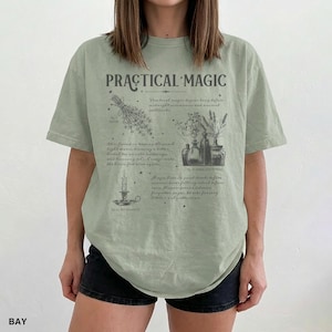 May include: Sage green t-shirt with the words "PRACTICAL MAGIC" in a vintage font. The shirt features botanical illustrations and text about spells and herbs. The shirt is a relaxed fit.