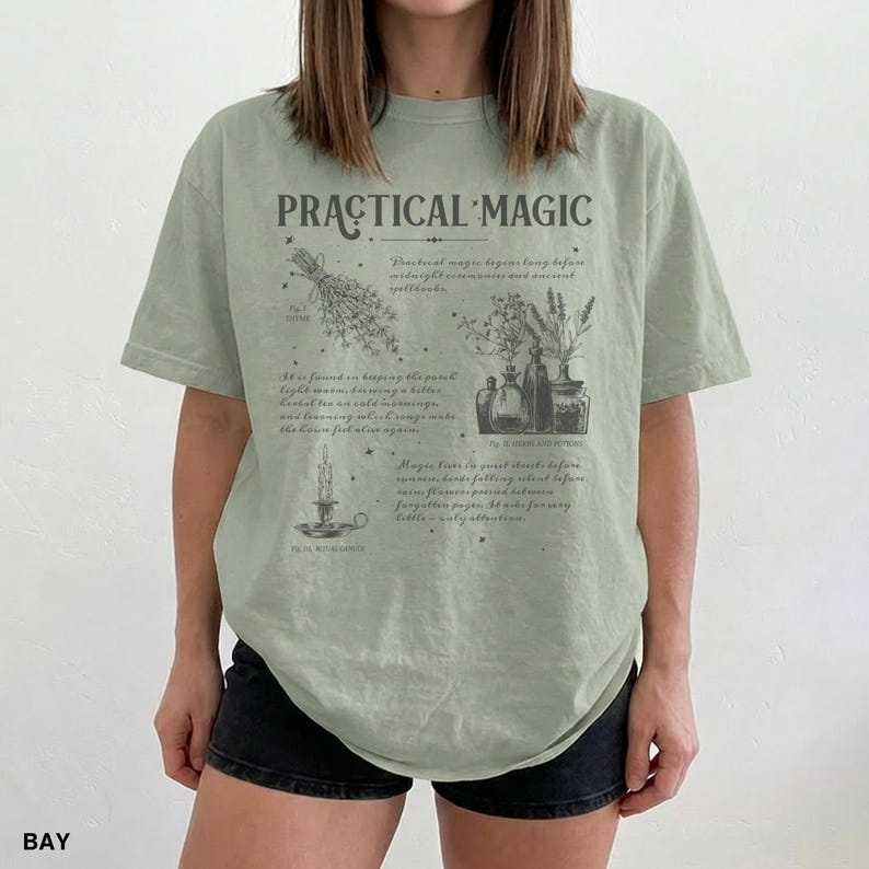 May include: Sage green t-shirt with the words "PRACTICAL MAGIC" in a vintage font. The shirt features botanical illustrations and text about spells and herbs. The shirt is a relaxed fit.