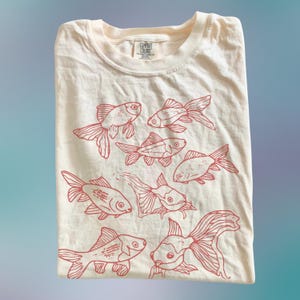 May include: A cream-colored t-shirt featuring a red line-art graphic print of seven goldfish swimming in various directions. The shirt has a crew neckline and a Comfort Colors brand label printed on the inside of the collar area.