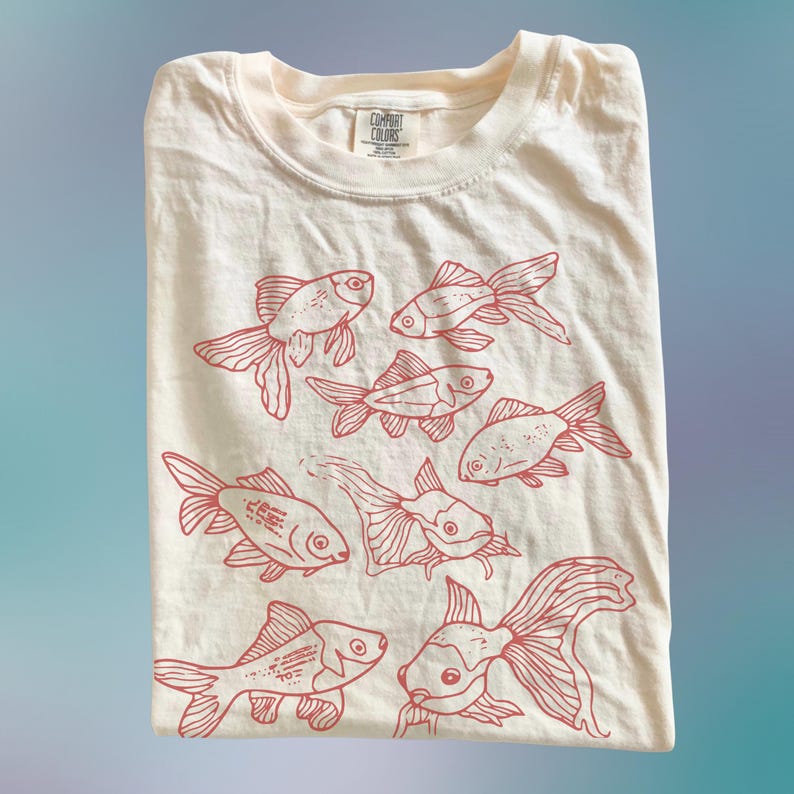 May include: A cream-colored t-shirt featuring a red line-art graphic print of seven goldfish swimming in various directions. The shirt has a crew neckline and a Comfort Colors brand label printed on the inside of the collar area.