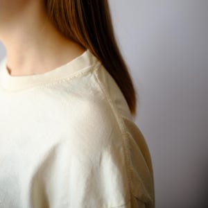 May include: A cream-colored crew neck t-shirt featuring a ribbed collar and visible shoulder seams. The fabric has a smooth texture and a light, off-white tone. The garment is shown from the side, highlighting the neckline and sleeve construction.