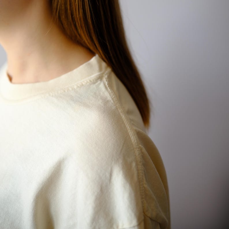 May include: A cream-colored crew neck t-shirt featuring a ribbed collar and visible shoulder seams. The fabric has a smooth texture and a light, off-white tone. The garment is shown from the side, highlighting the neckline and sleeve construction.