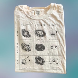 Puede incluir: Camiseta de color crema con un gráfico que presenta una cuadrícula de fenómenos cósmicos, incluyendo un agujero negro y una nebulosa. La camiseta tiene las palabras "Caótico, Neutral, Legal" y "Mal, Neutral, Bueno" impresas.