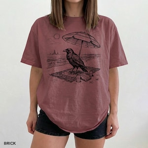 May include: Brick-colored t-shirt featuring a black graphic of a raven on a beach, under a striped umbrella. The design includes a sun, waves, and a small bottle. The word "BRICK" is printed at the bottom.