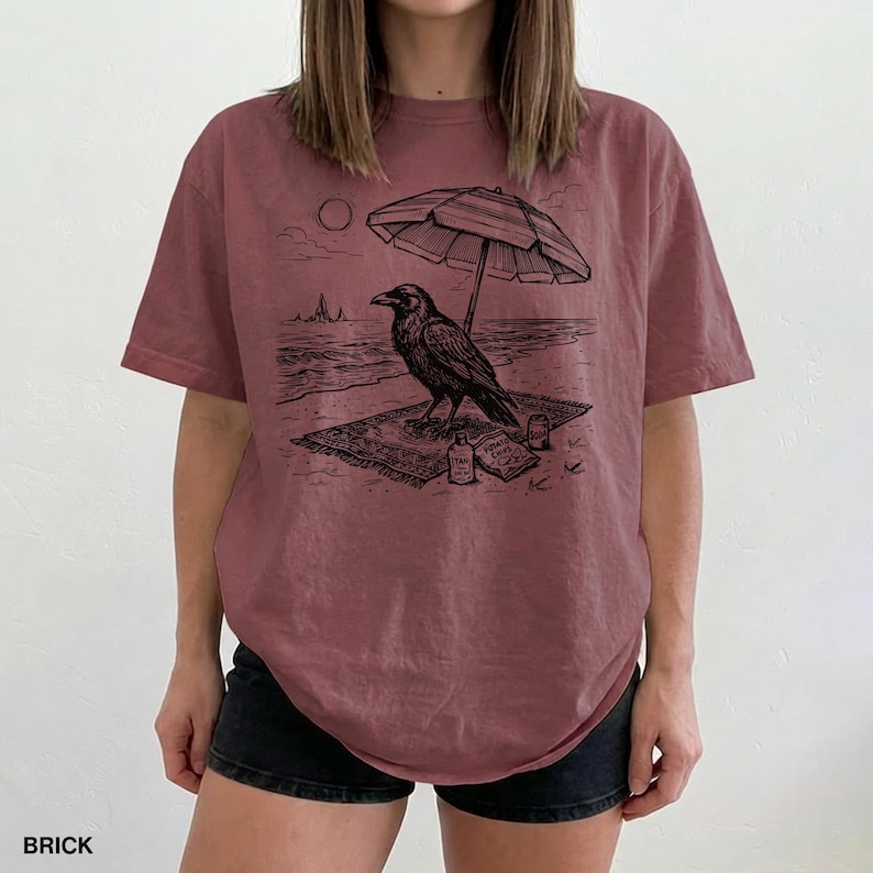 May include: Brick-colored t-shirt featuring a black graphic of a raven on a beach, under a striped umbrella. The design includes a sun, waves, and a small bottle. The word "BRICK" is printed at the bottom.