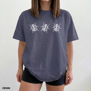 May include: A slate blue short-sleeve t-shirt featuring a white graphic print of three ladybugs arranged in a horizontal row across the chest. The shirt has a crew neckline and a relaxed fit, showcasing a simple insect illustration design.