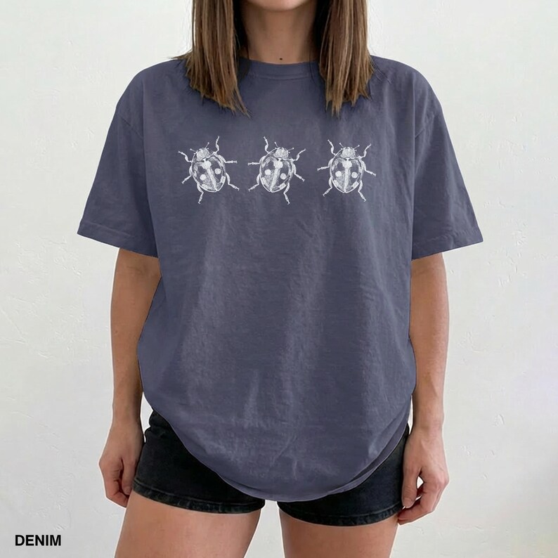 May include: A slate blue short-sleeve t-shirt featuring a white graphic print of three ladybugs arranged in a horizontal row across the chest. The shirt has a crew neckline and a relaxed fit, showcasing a simple insect illustration design.