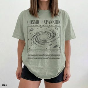 May include: A light green t-shirt with the text "COSMIC EXPANSION" and a graphic of a galaxy. The shirt also has text describing the expansion of the universe. The shirt is worn by a person.