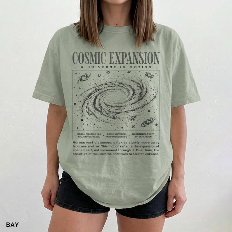 May include: A light green t-shirt with the text "COSMIC EXPANSION" and a graphic of a galaxy. The shirt also has text describing the expansion of the universe. The shirt is worn by a person.