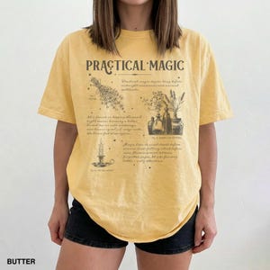 May include: A yellow t-shirt with the words "PRACTICAL MAGIC" in a vintage font. The shirt features illustrations of herbs, potions, and a candle. The word "BUTTER" is printed at the bottom.